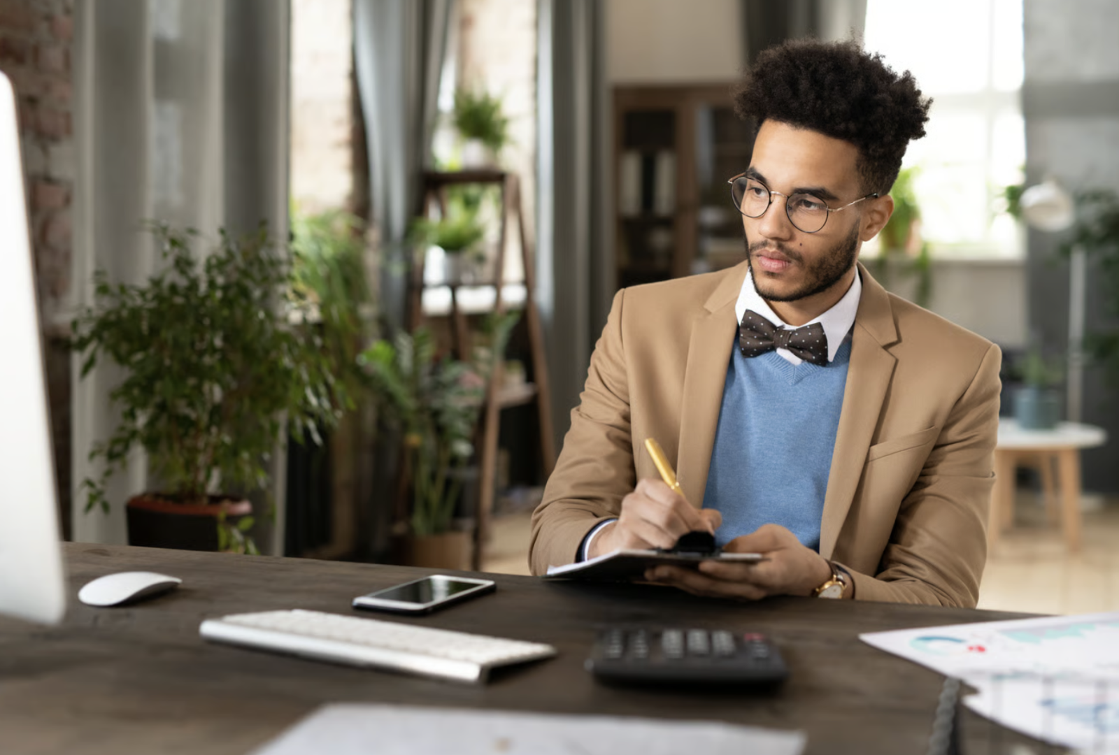 An accountant making notes with a calculator, keyboard and financial paperwork spread across the desk